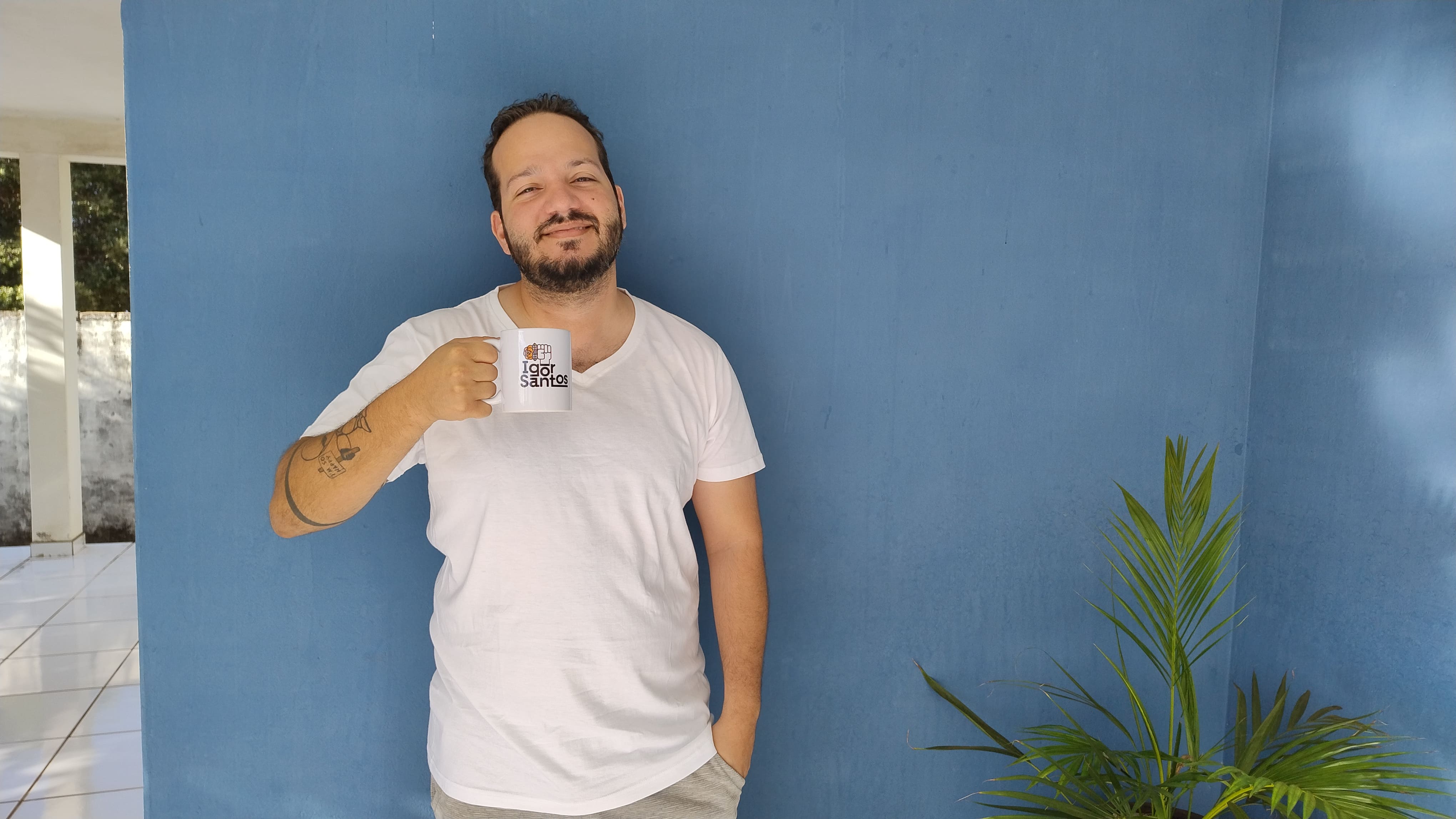 Igor de camiseta branca segurando uma caneca com a logo "Igor Santos", azul, com uma flor laranja que sai do "o" de Igor. A foto é despojada, com Igor sorrindo e bebendo café encostado na parede. Ao lado esquerdo, uma planta que não sei dizer qual é.